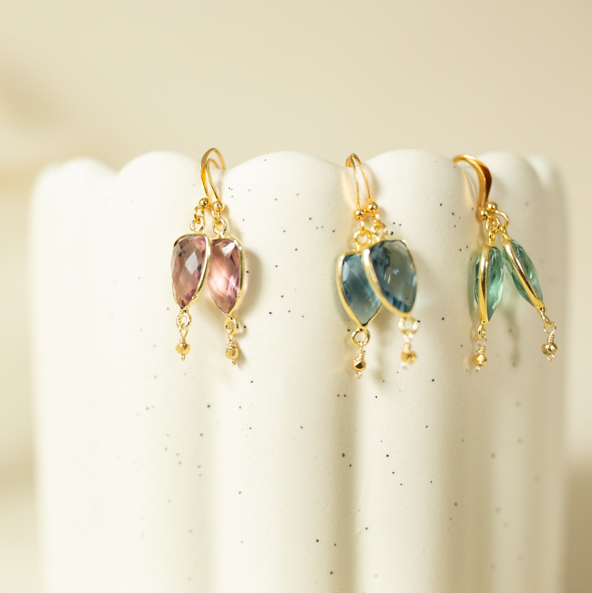Three pairs of gold dangle earrings with marquise-cut gemstones in pink, blue, and green, displayed on the scalloped edge of a white speckled ceramic pedestal.