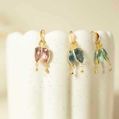 Three pairs of gold dangle earrings with marquise-cut gemstones in pink, blue, and green, displayed on the scalloped edge of a white speckled ceramic pedestal.