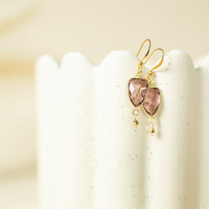 A pair of gold drop earrings with heart-shaped pinkish-brown stones, displayed on a white speckled ceramic stand against a warm beige background.