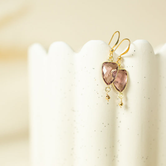 A pair of gold drop earrings with heart-shaped pinkish-brown stones, displayed on a white speckled ceramic stand against a warm beige background.