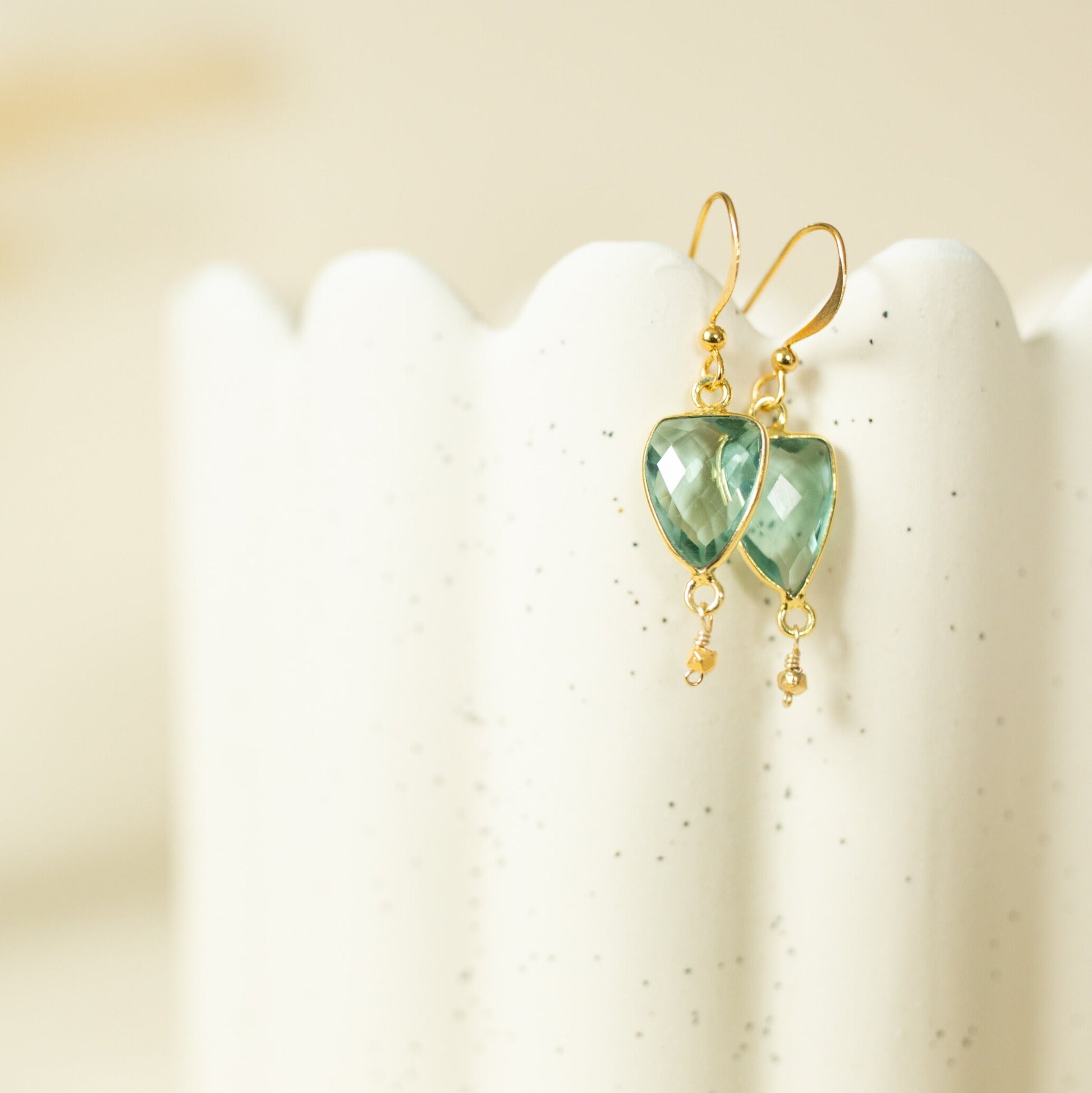 A pair of gold drop earrings with faceted light green heart-shaped gemstones and small dangling gold beads, displayed on a white scalloped stand against a soft beige background.