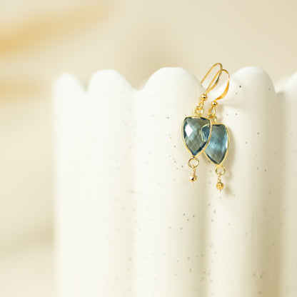 A pair of gold drop earrings with heart-shaped blue-green stones and small dangling beads, displayed on a white speckled stand against a soft beige background.