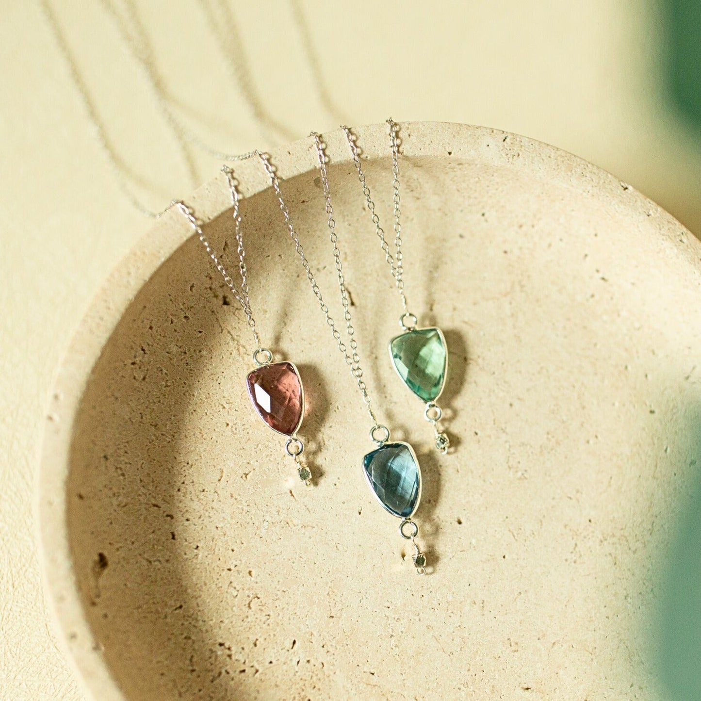 Three silver pendant necklaces with marquise-cut gemstones in pink, green, and blue, displayed on a textured beige ceramic dish.