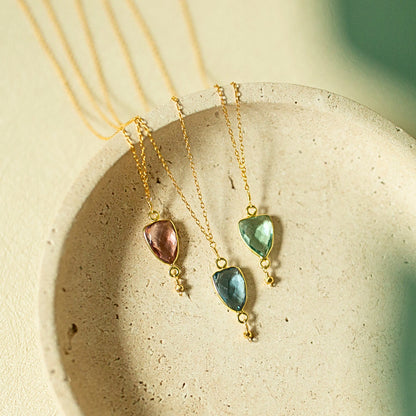 Three gold chain necklaces with teardrop-shaped gemstone pendants in pink, blue, and green, displayed on a textured beige ceramic dish.