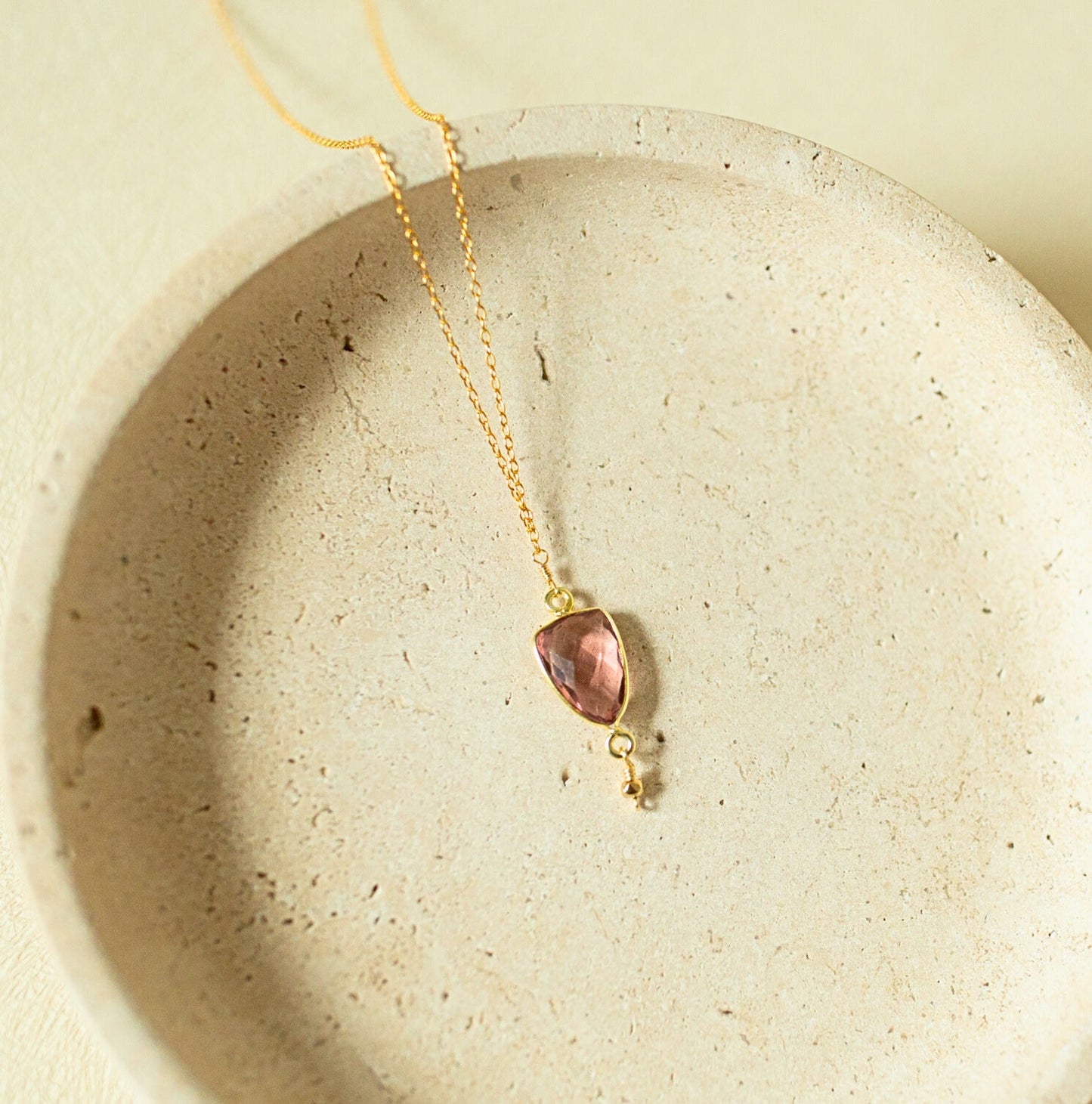 A delicate gold chain necklace with a small teardrop-shaped pink gemstone pendant resting on a textured beige ceramic dish.
