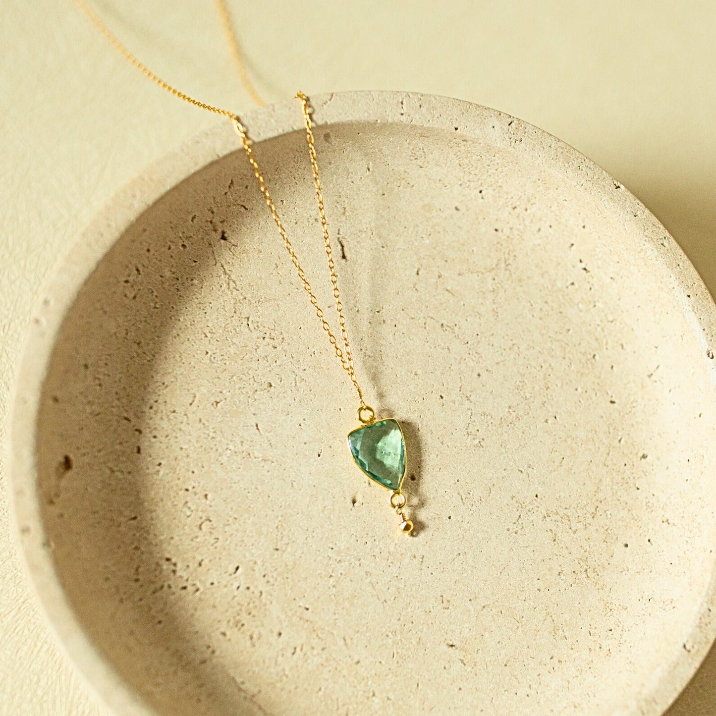 A delicate gold chain necklace with a small, irregular-cut green gemstone pendant, displayed inside a textured beige ceramic dish.