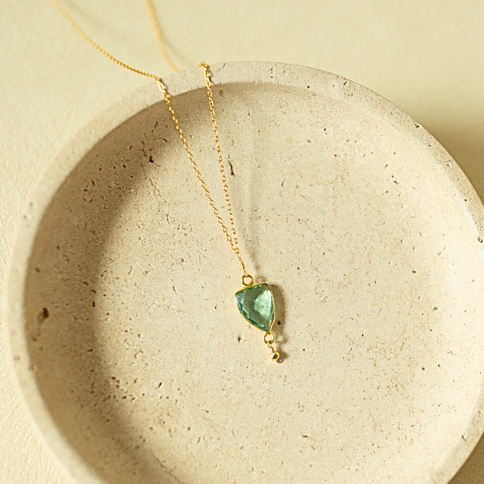 A delicate gold chain necklace with a small, irregular-cut green gemstone pendant, displayed inside a textured beige ceramic dish.