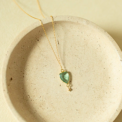 A delicate gold chain necklace with a small, irregular-cut green gemstone pendant, displayed inside a textured beige ceramic dish.
