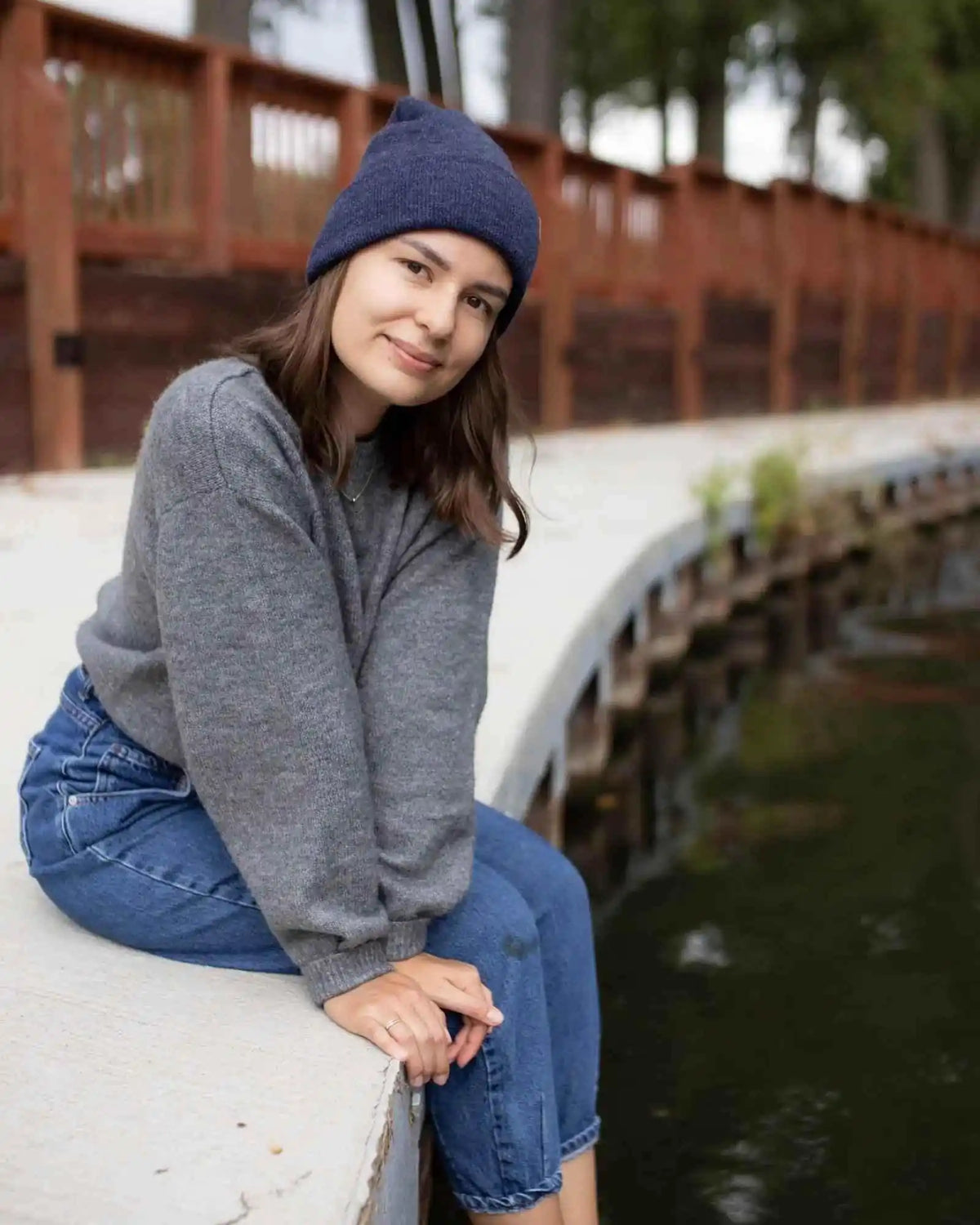 Woman sitting by a water body wearing a gray sweater and blue beanie