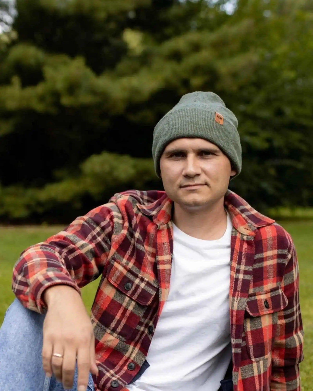 Person wearing a plaid shirt and beanie sitting outdoors with trees in the background