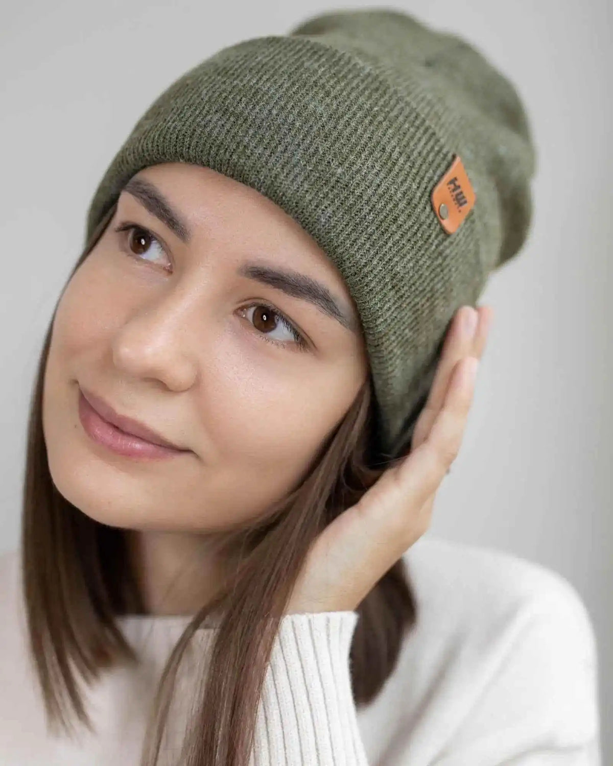 Woman wearing a green knit beanie with a brand logo on a plain background