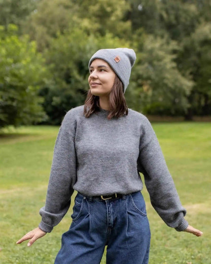 Person wearing a gray sweater and beanie standing outdoors with greenery in the background