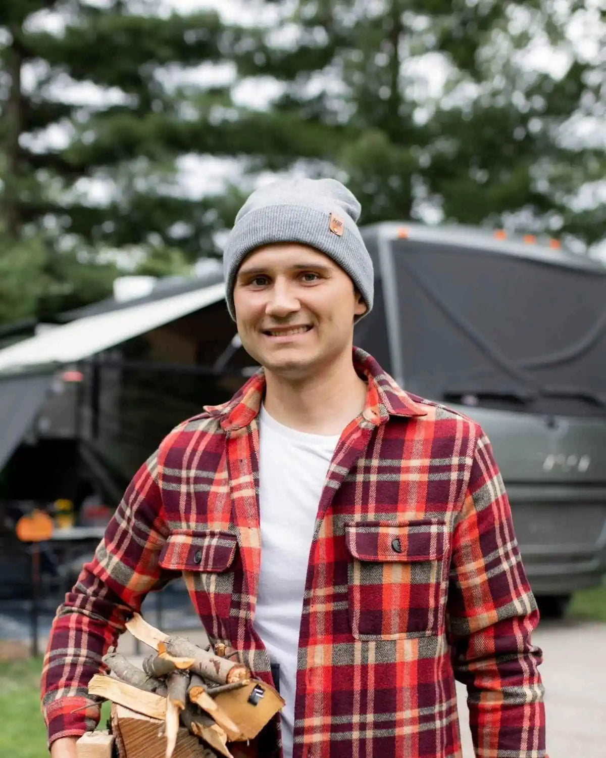 Man in a plaid shirt and beanie holding firewood with a trailer in the background