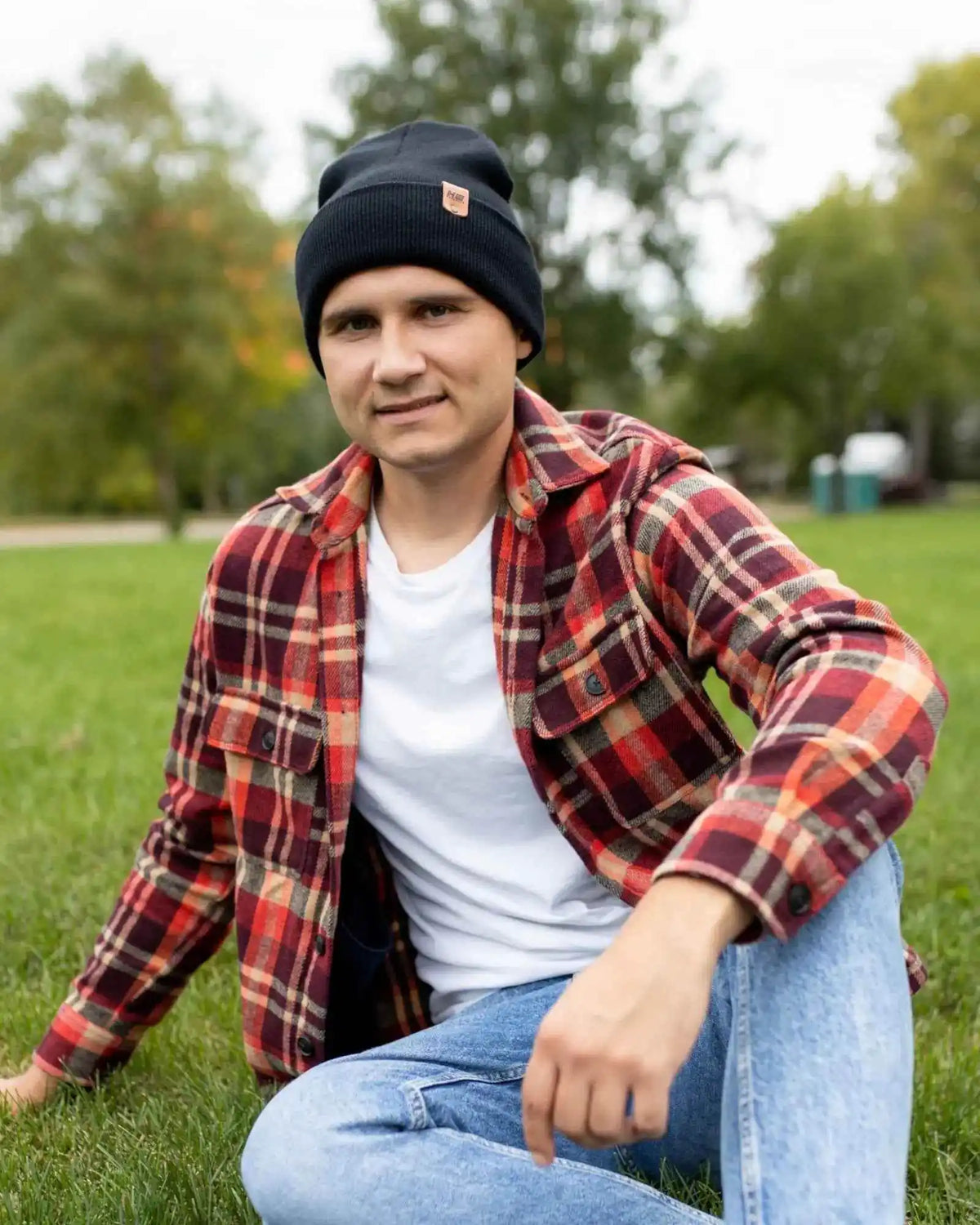 Person wearing a red plaid shirt, white t-shirt, and black beanie sitting on grass with trees in the background