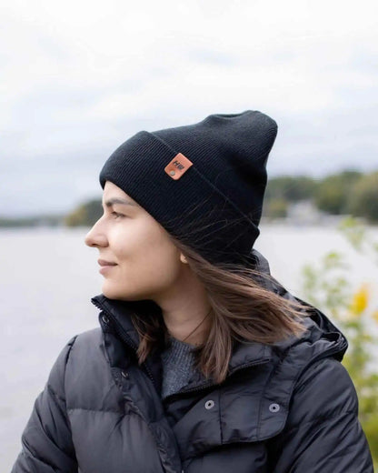 Person wearing a black beanie with a logo, standing by a body of water.