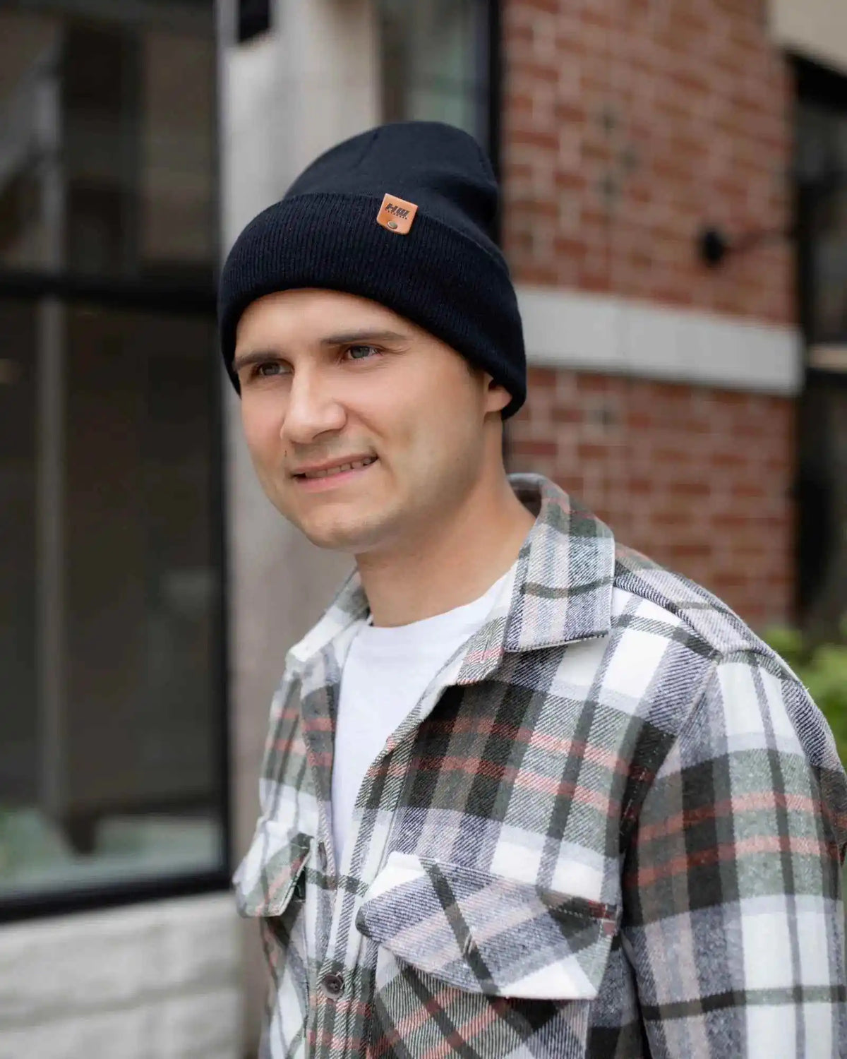 Man wearing a black beanie and plaid shirt standing outdoors with a brick building in the background
