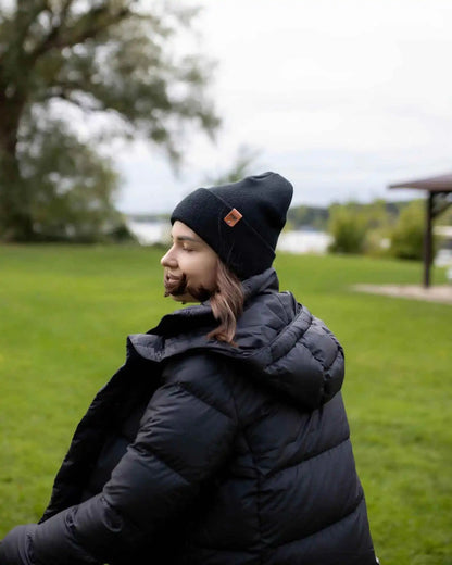 Person wearing a black beanie and puffer jacket sitting on grass with trees and a lake in the background