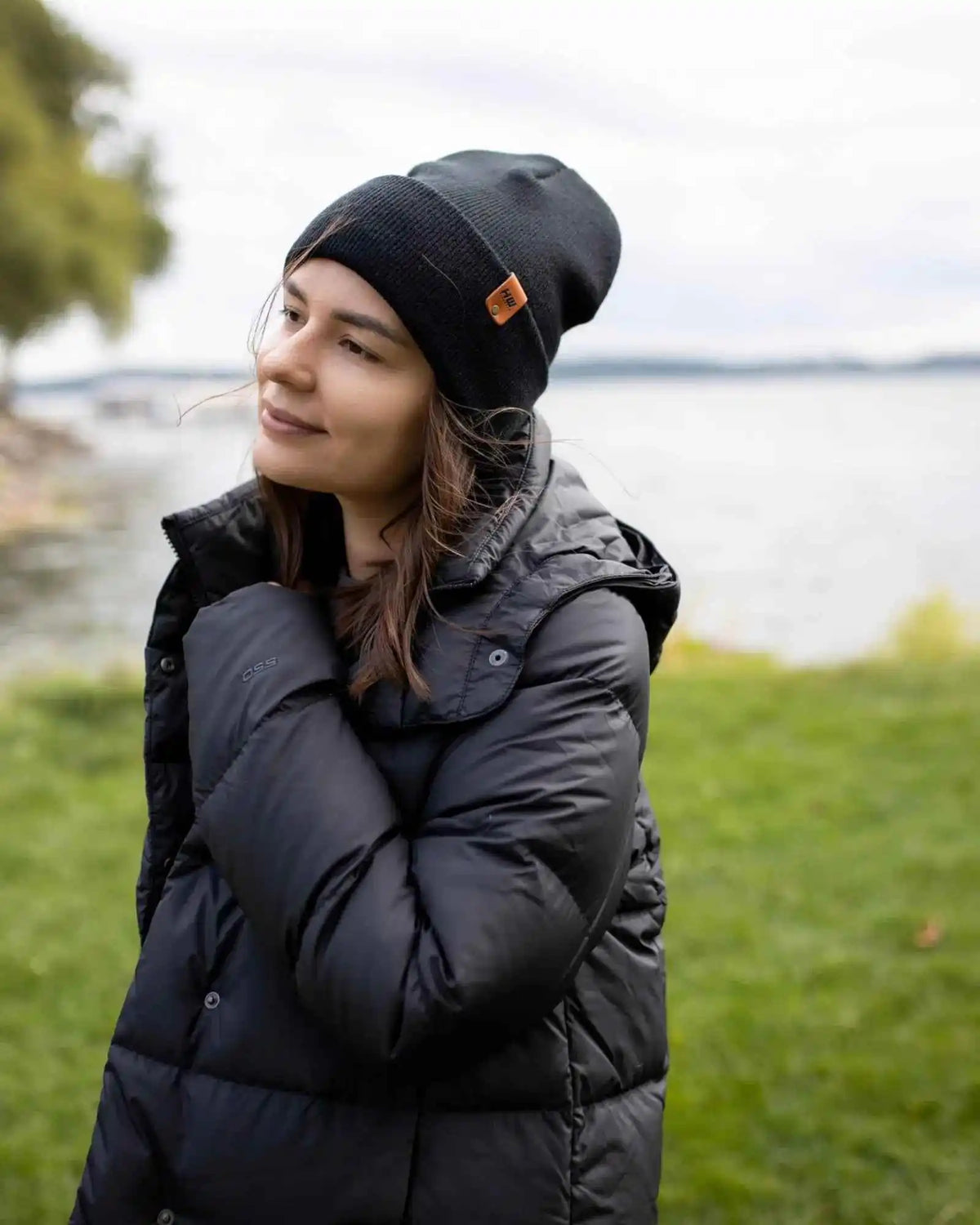 Person wearing a black puffer jacket and beanie with a logo, standing outdoors by a body of water.