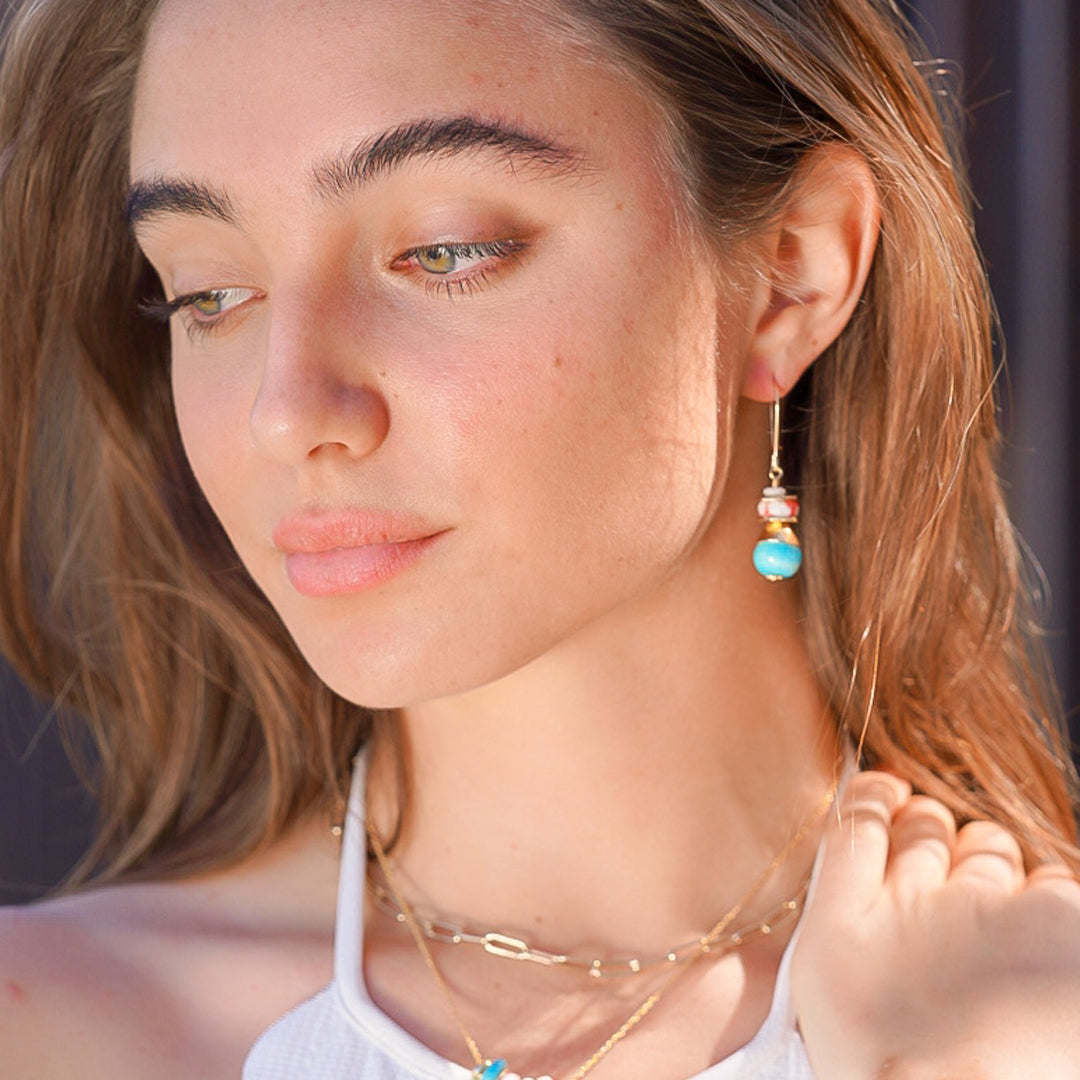 A close-up lifestyle shot of a woman wearing a delicate gold dangle earring with a light blue bead and a small gold bead accent, paired with layered gold necklaces.