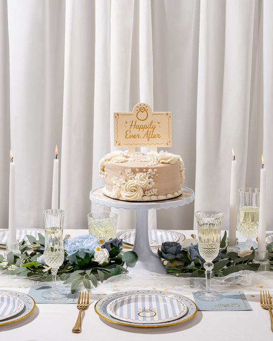 Wedding cake with 'Happily Ever After' topper on a table set with plates, glasses, and flowers.