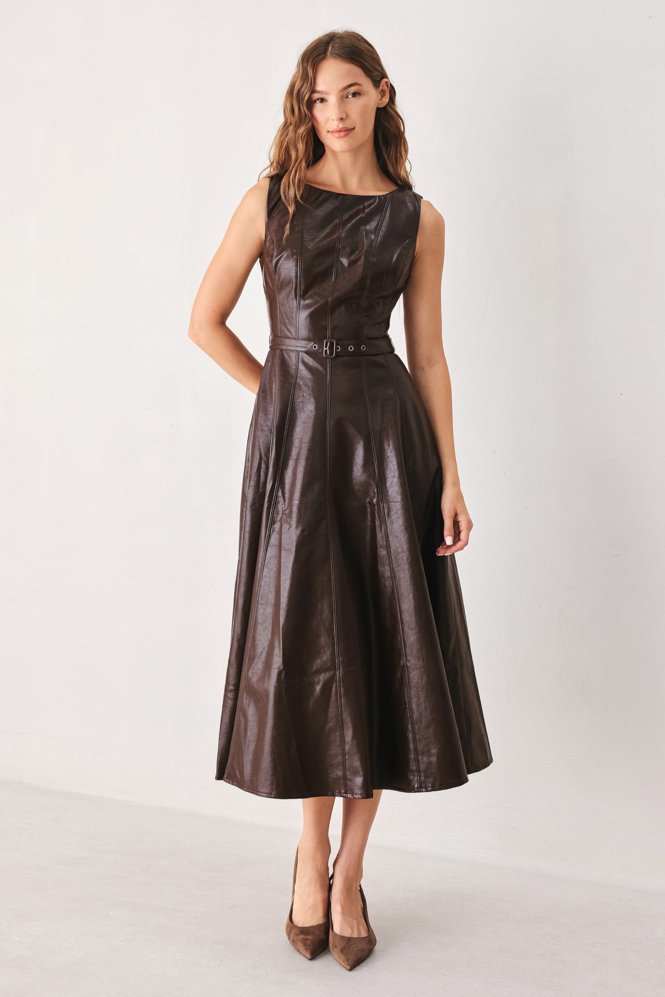 A woman models a sleeveless, dark brown faux-leather midi dress with a defined waist and a flared skirt, paired with brown pointed-toe heels, against a plain light gray studio background.