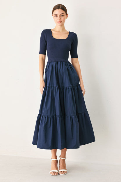 A woman modeling a navy blue midi dress with a square neckline, short sleeves, and a tiered skirt, paired with white heeled sandals against a plain light gray studio background.