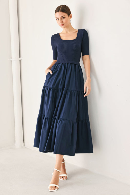 Navy blue midi dress with a ribbed short-sleeve bodice and a tiered fabric skirt, worn by a woman leaning against a wall.