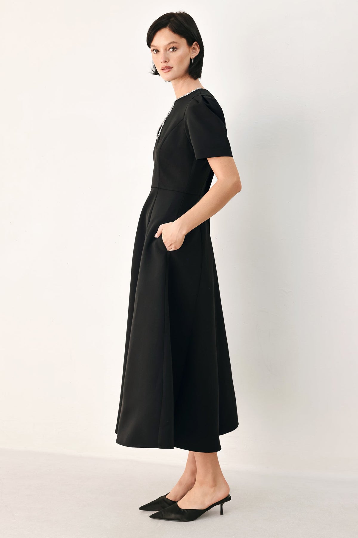 Side view of the black midi dress showing a short puff sleeve and flared skirt, with the model’s hand in a side pocket.