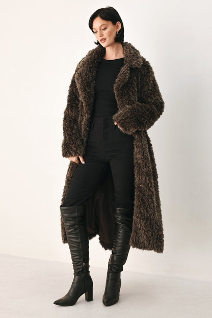 A model wearing a long, dark brown shaggy faux fur coat over a black outfit with black leather knee-high boots, photographed against a plain light gray studio background.