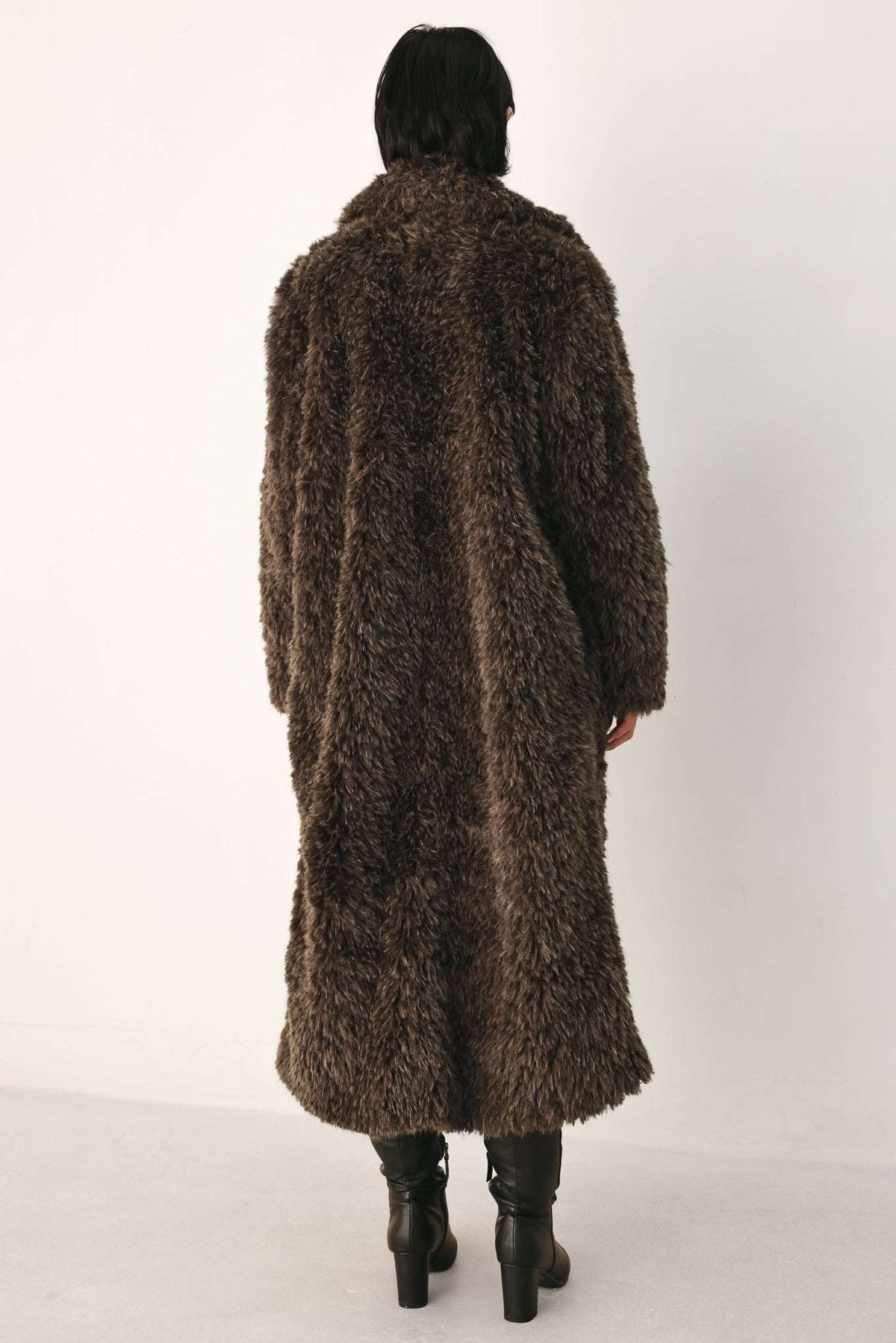 A person viewed from the back wearing a long, shaggy brown faux-fur coat over black knee-high boots, standing against a plain light background.