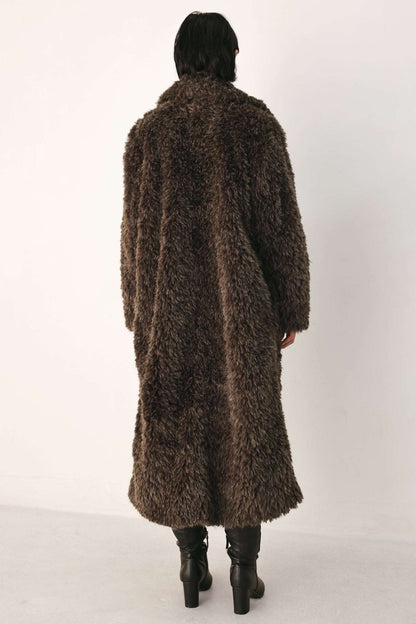 A person viewed from the back wearing a long, shaggy brown faux-fur coat over black knee-high boots, standing against a plain light background.