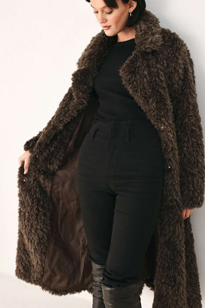 A woman wearing a long, dark brown shaggy faux-fur coat over a black ribbed top, black pants, and black boots, standing against a plain white background.