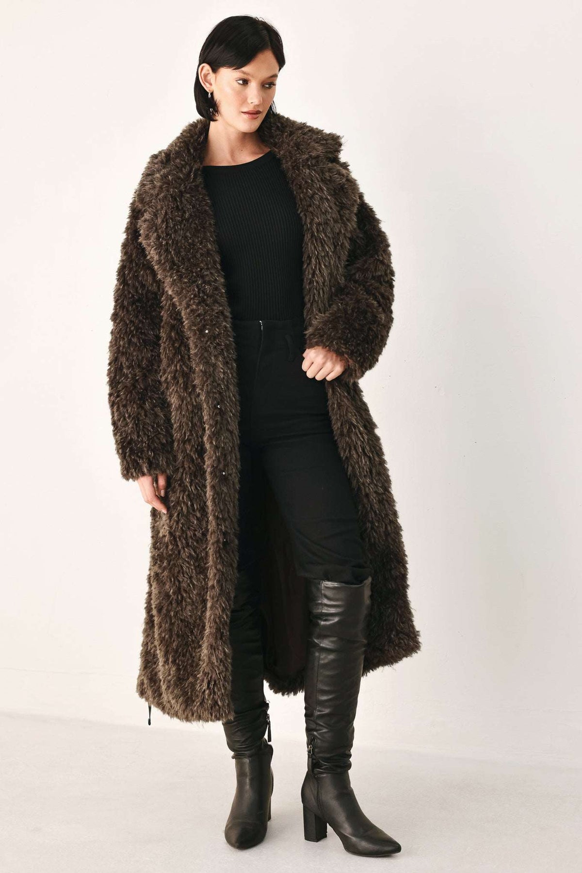 A woman modeling a long, dark brown shaggy faux-fur coat over a black outfit and black knee-high boots, photographed against a plain light studio background.