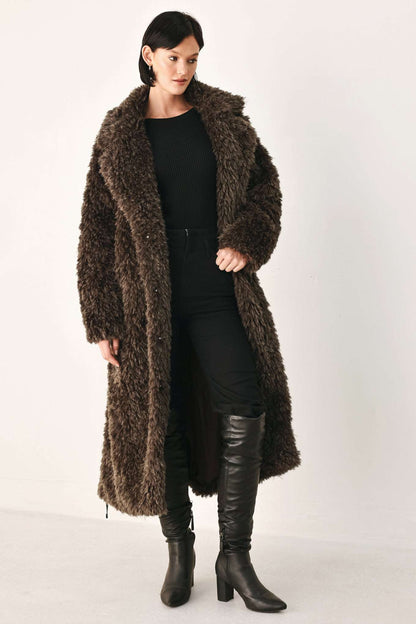 A woman modeling a long, dark brown shaggy faux-fur coat over a black outfit and black knee-high boots, photographed against a plain light studio background.