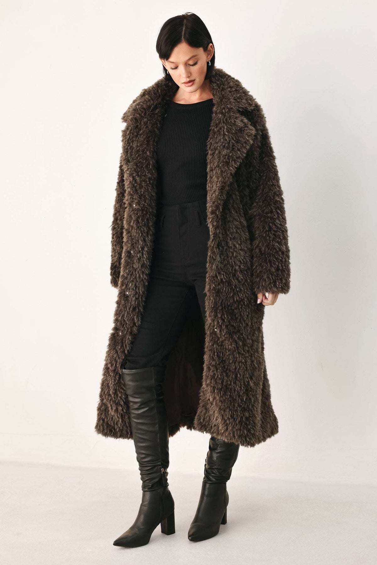 A model wearing a long, dark brown shaggy faux fur coat over a black outfit and black knee-high boots, standing against a plain off-white studio background.