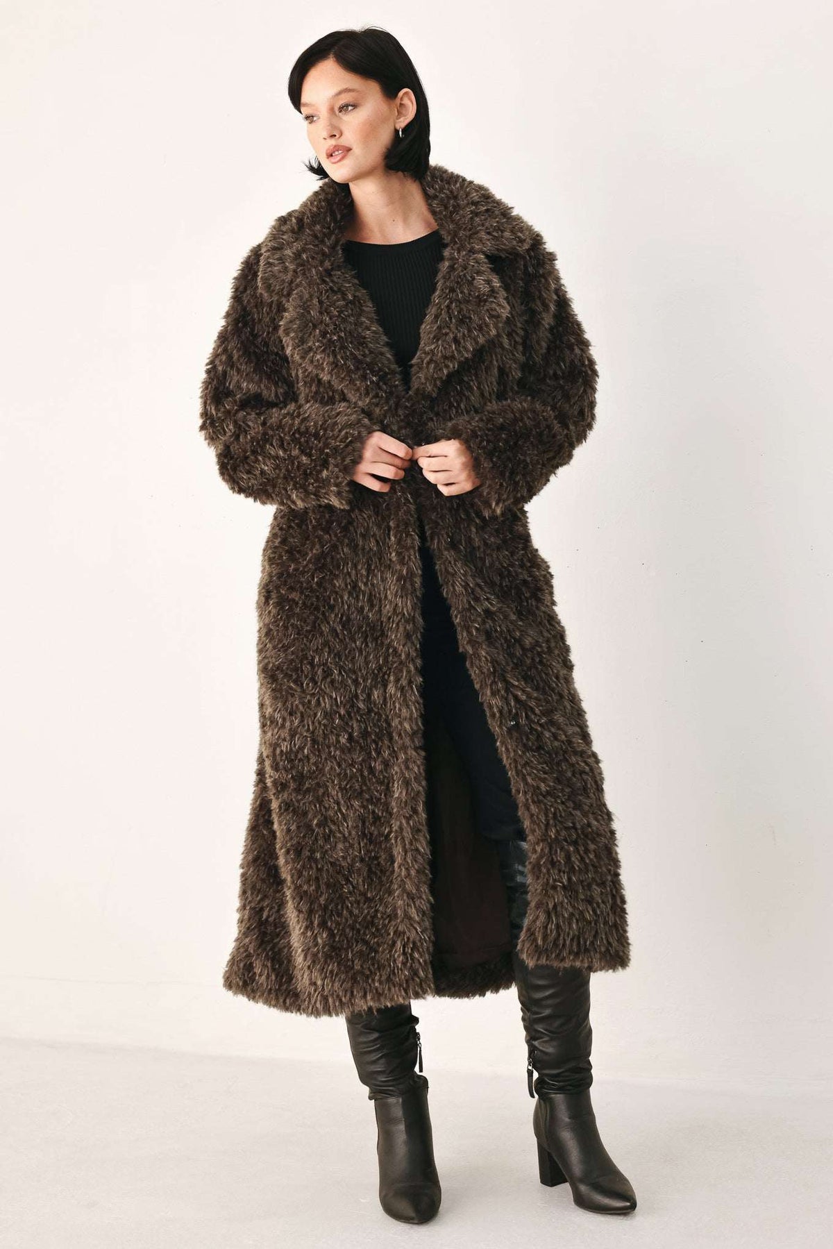 A woman modeling a long, shaggy brown faux fur coat over a black outfit and black knee-high boots, photographed against a plain light studio background.