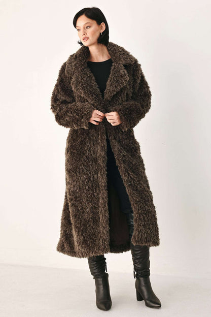 A woman modeling a long, shaggy brown faux fur coat over a black outfit and black knee-high boots, photographed against a plain light studio background.