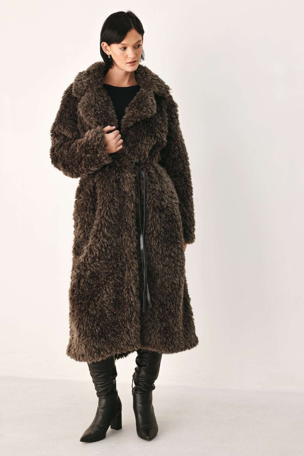 A model wearing a long, dark brown shaggy faux fur coat over a black top and black leather boots, standing against a plain off-white studio background.