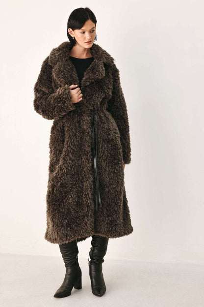 A model wearing a long, dark brown shaggy faux fur coat over a black top and black leather boots, standing against a plain off-white studio background.