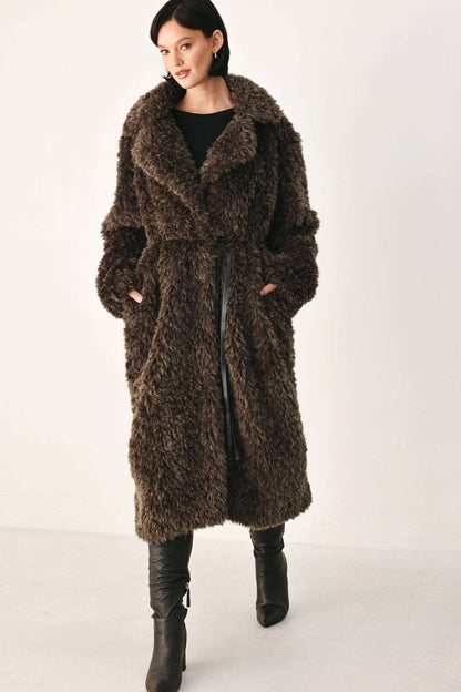 A woman modeling a long, shaggy brown faux fur coat with a wide collar, worn over a black top and black knee-high boots, against a plain white studio background.