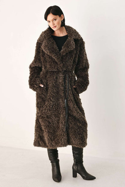 A woman modeling a long, shaggy brown faux fur coat with a black top and black heeled boots, standing against a plain light gray studio background.
