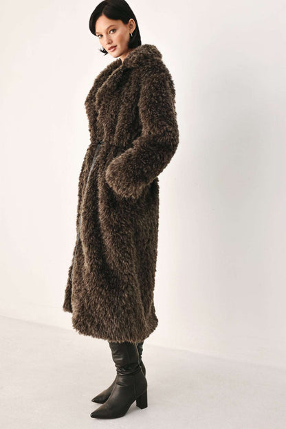 A woman modeling a long, shaggy brown faux fur coat with black leather ankle boots, standing against a plain off-white studio background.