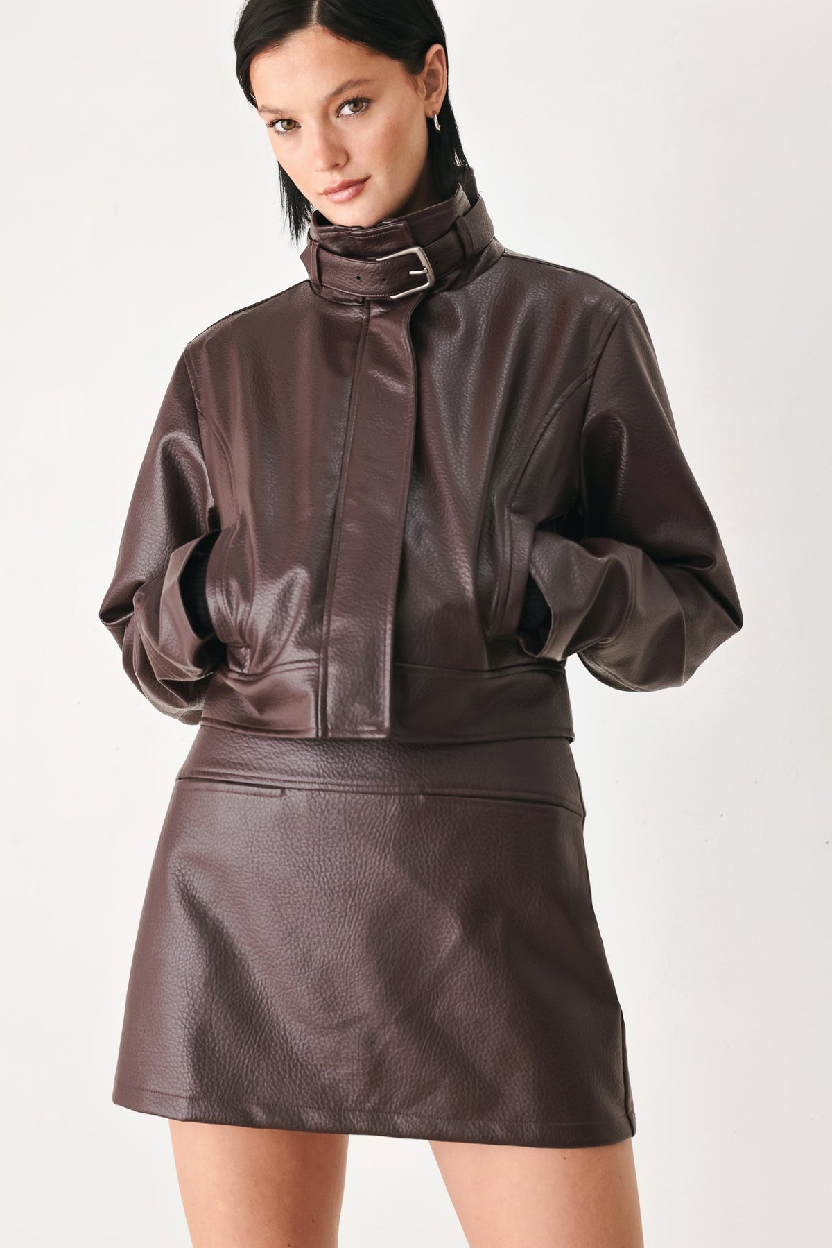 A model wearing a dark brown faux-leather jacket and matching mini skirt against a plain light background.