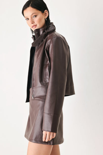 A model wearing a dark brown faux-leather jacket and matching mini skirt set, photographed against a clean off-white studio background.