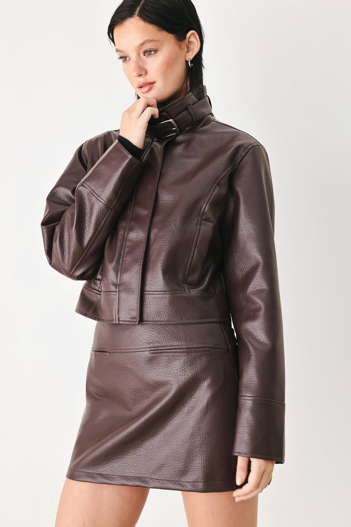 A model wearing a dark brown faux leather jacket and matching mini skirt set, photographed against a clean light gray studio background.