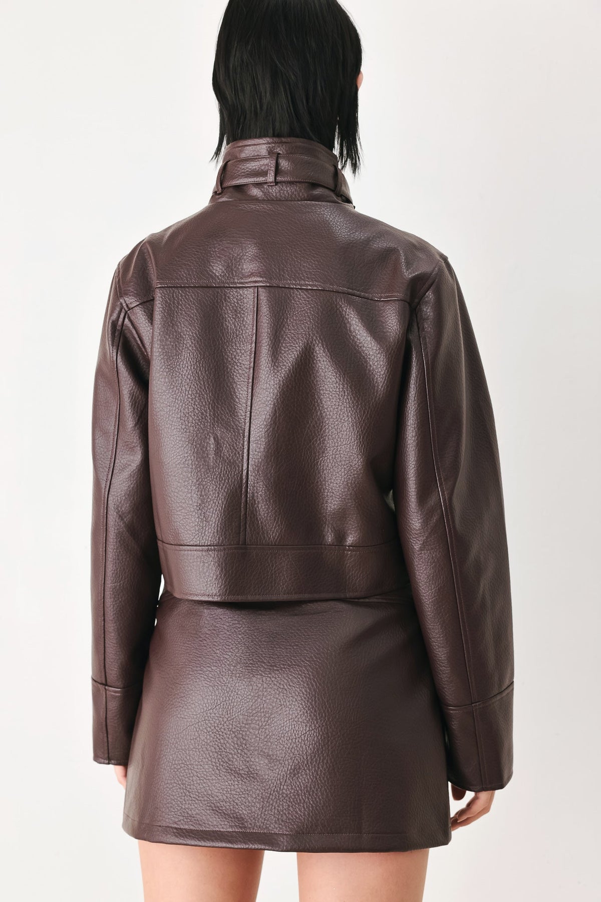 A person shown from the back wearing a dark brown textured faux-leather jacket and matching mini skirt against a plain light background.