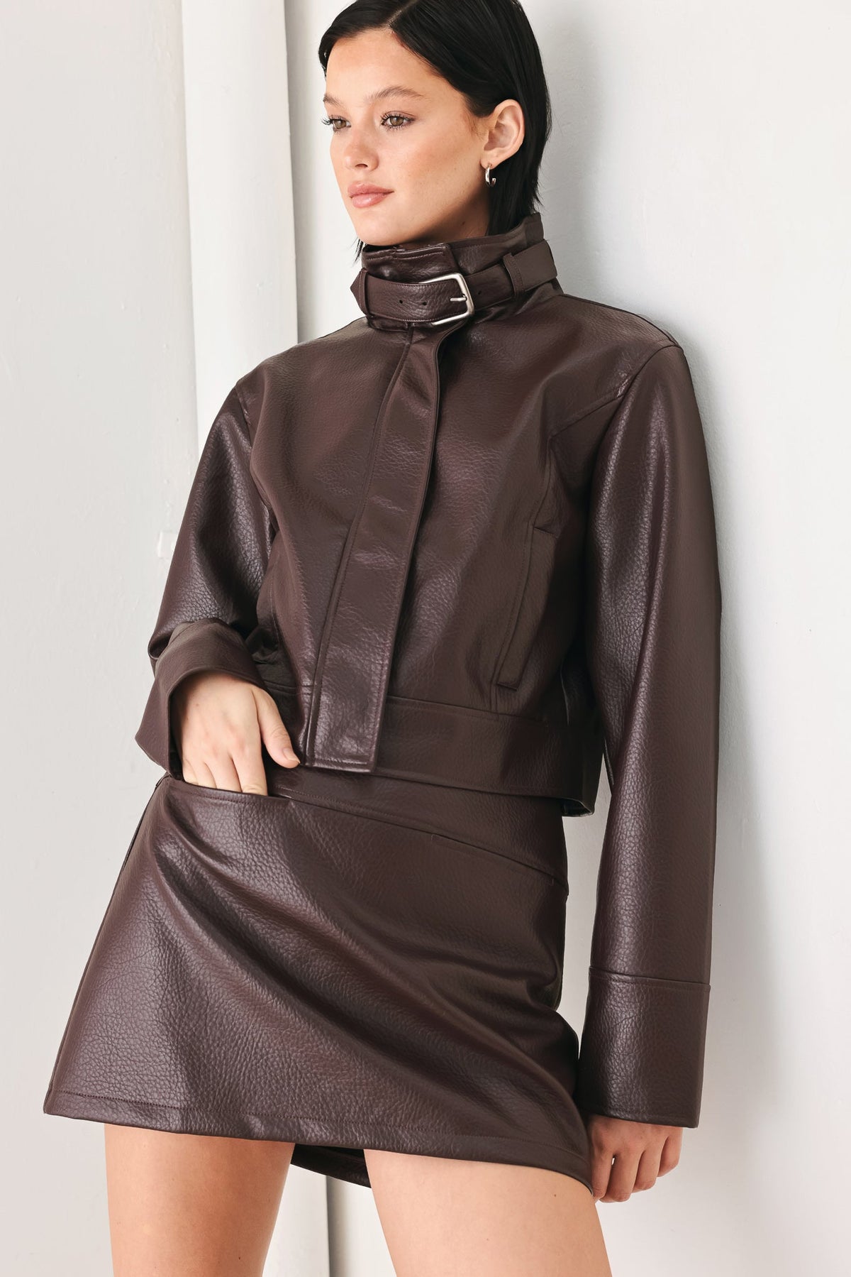 A model wearing a dark brown faux leather two-piece outfit consisting of a cropped long-sleeve jacket with a high collar and matching mini skirt, posed against a light gray wall.