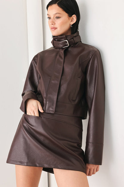A model wearing a dark brown faux leather two-piece outfit consisting of a cropped long-sleeve jacket with a high collar and matching mini skirt, posed against a light gray wall.