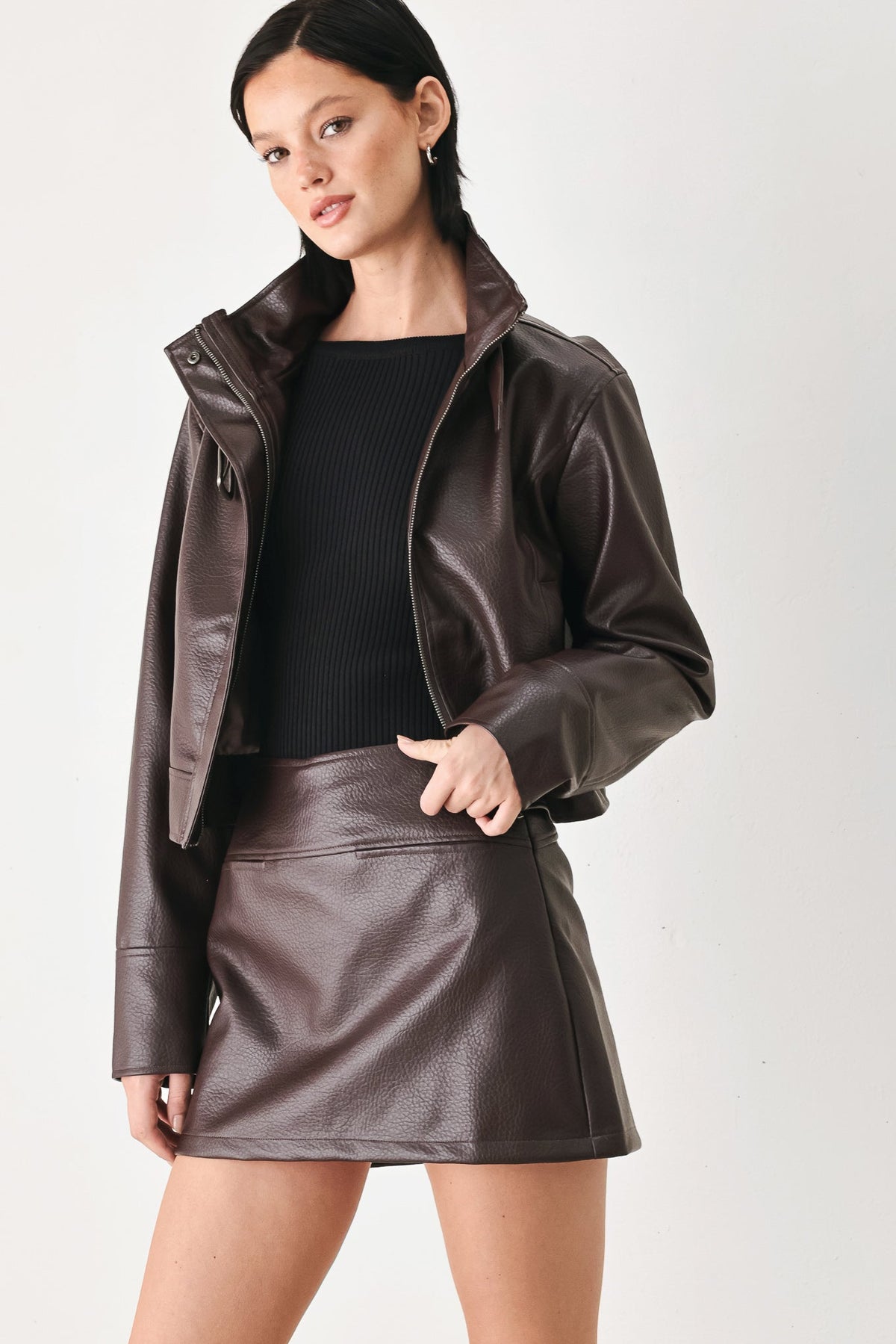 A woman modeling a dark brown faux-leather jacket and matching mini skirt over a black ribbed top, photographed against a plain light gray studio background.