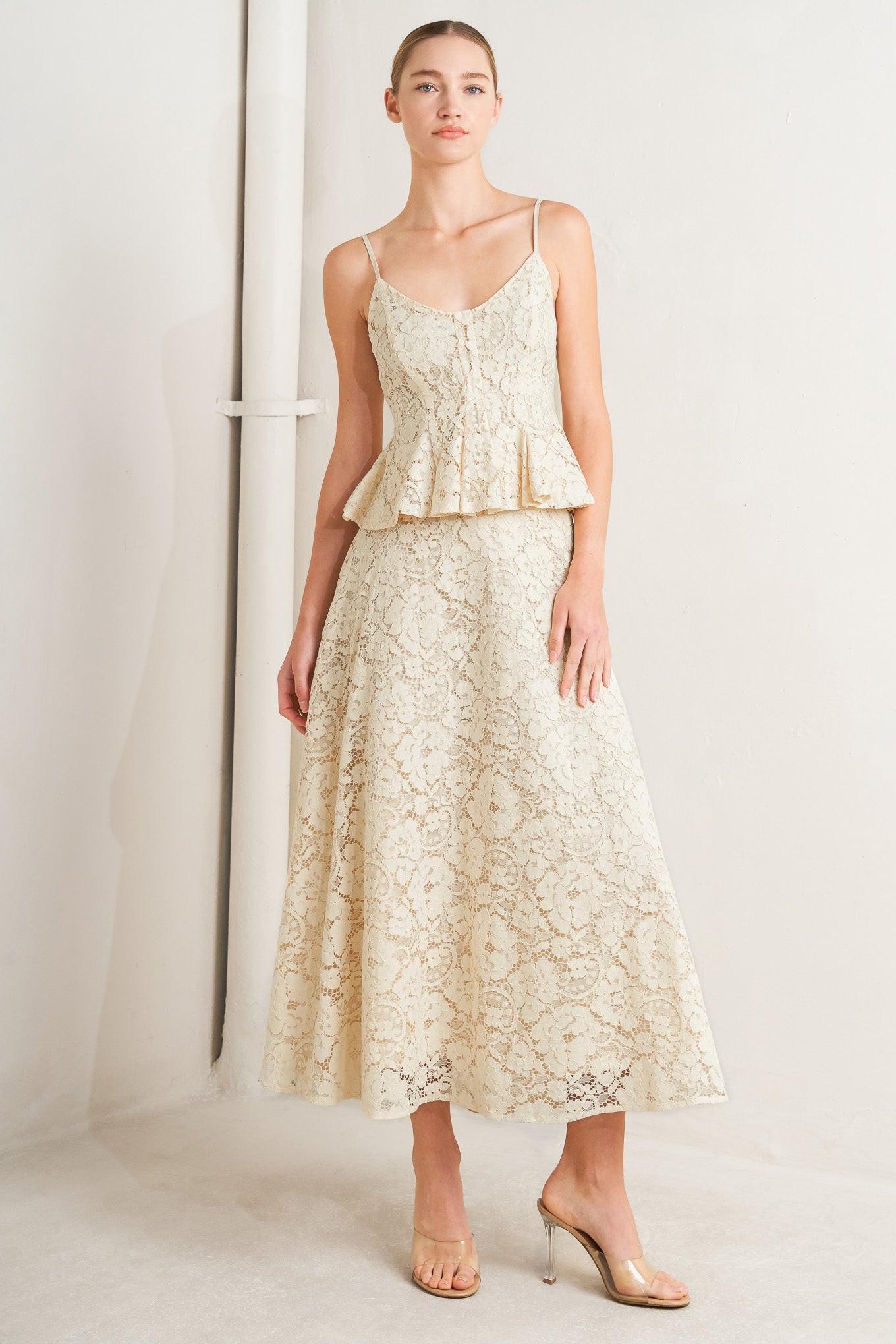 A model wears a matching cream lace camisole top and midi skirt set, styled with clear high-heeled sandals against a minimalist studio backdrop.